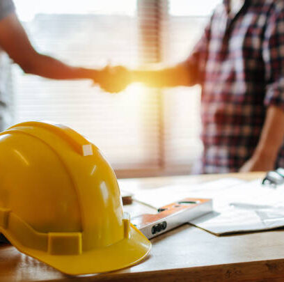 yellow safety helmet on workplace desk with construction worker team hands shaking greeting start up plan new project contract in office center at construction site, partnership and contractor concept